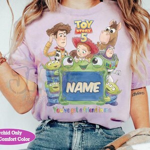 May include: A light purple t-shirt featuring a Toy Story 5 graphic with Woody, Jessie, Bullseye, Buzz Lightyear, and three aliens surrounding a green frog holding a blue sign that says NAME, with the text You've got a friend in me below.