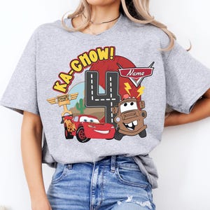 May include: A light gray t-shirt featuring a colorful graphic design from the movie Cars. The design includes the phrase "Ka-Chow!", the number 4, and images of Lightning McQueen and Mater. The shirt is a crew neck.