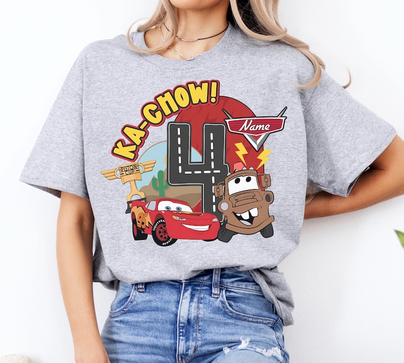 May include: A light gray t-shirt featuring a colorful graphic design from the movie Cars. The design includes the phrase "Ka-Chow!", the number 4, and images of Lightning McQueen and Mater. The shirt is a crew neck.