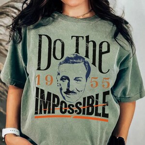 Pu&ograve; includere: T-shirt verde salvia con la scritta "Do The Impossible" in nero, con l'immagine del volto di un uomo al centro. I numeri "1955" sono arancioni. La maglietta &egrave; in stile vintage.