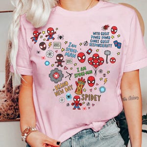 Puede incluir: Camiseta rosa claro con un dise&ntilde;o en forma de coraz&oacute;n de Spider-Man. El dise&ntilde;o incluye los textos "I AM SPIDER-MAN", "SPIDEY" y "BRAND NEW DAY", junto con estrellas, rayos y otros elementos de c&oacute;mic.