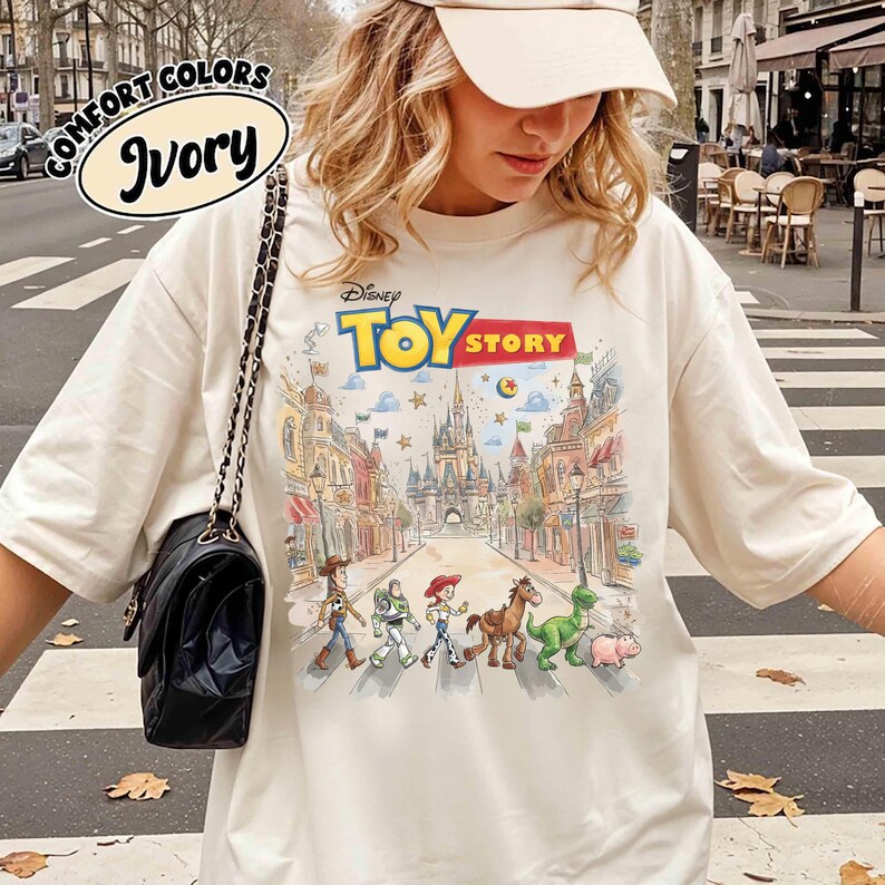 May include: Ivory-colored t-shirt featuring the Disney Toy Story logo and a colorful graphic of the characters crossing a street in front of a castle. The shirt has the words "Comfort Colors" above the word "Ivory".