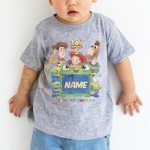 May include: A heather grey short-sleeved t-shirt featuring a colourful Toy Story 5 graphic with characters including Woody, Jessie, Buzz Lightyear, and aliens. The design includes a central green frame with the text NAME and You've got a friend in me.