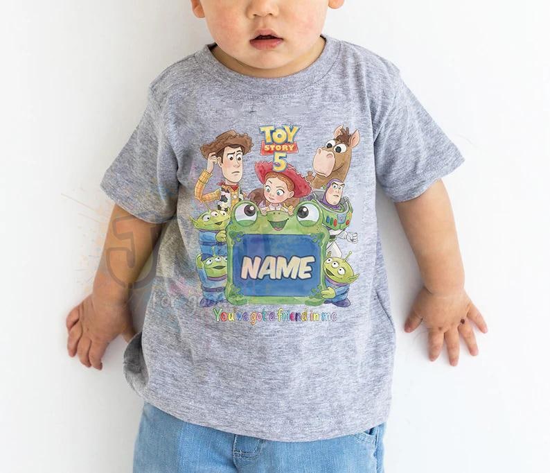 May include: A heather grey short-sleeved t-shirt featuring a colourful Toy Story 5 graphic with characters including Woody, Jessie, Buzz Lightyear, and aliens. The design includes a central green frame with the text NAME and You've got a friend in me.