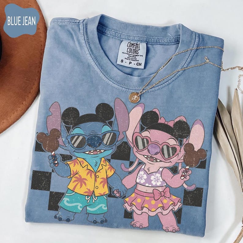 May include: Blue jean colored t-shirt featuring a graphic of Stitch and Angel from Lilo & Stitch, each holding a Mickey Mouse-shaped ice cream bar. Stitch is wearing sunglasses, a Hawaiian shirt, and shorts. Angel is wearing sunglasses and a floral bikini. A gold necklace is draped across the shirt.
