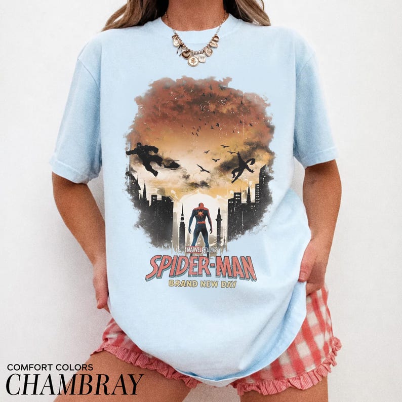 Pu&ograve; includere: Una t-shirt azzurra con una stampa grafica di Spider-Man in piedi davanti alla sagoma di una citt&agrave; su un cielo al tramonto. Il design include sagome di figure volanti, uccelli e la scritta Spider-Man Brand New Day in lettere rosse e bianche.