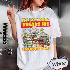 May include: A white short-sleeve t-shirt featuring a colorful graphic print of various cartoon toys and the text MY BOY ONLY BREAKS HIS FAVORITE TOYS in yellow and red lettering. The design includes a doll, a jack-in-the-box, and other figures.