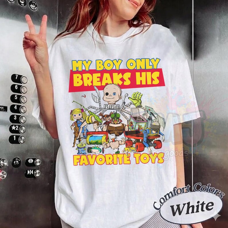 May include: A white short-sleeve t-shirt featuring a colorful graphic print of various cartoon toys and the text MY BOY ONLY BREAKS HIS FAVORITE TOYS in yellow and red lettering. The design includes a doll, a jack-in-the-box, and other figures.