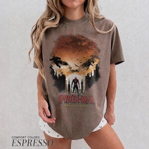 Pu&ograve; includere: Una t-shirt marrone a maniche corte con una stampa grafica di Spider-Man in piedi in una silhouette cittadina contro un cielo al tramonto. Il design include il testo Spider-Man e Brand New Day in lettere rosse e gialle con uccelli e figure.