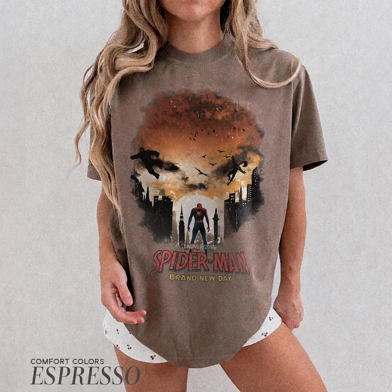 Pu&ograve; includere: Una t-shirt marrone a maniche corte con una stampa grafica di Spider-Man in piedi in una silhouette cittadina contro un cielo al tramonto. Il design include il testo Spider-Man e Brand New Day in lettere rosse e gialle con uccelli e figure.