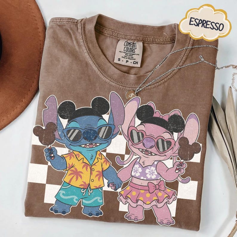 May include: Espresso-colored t-shirt featuring a cartoon graphic of two characters, one blue and one pink, holding ice cream. The blue character wears sunglasses and a Hawaiian shirt. The pink character wears heart-shaped sunglasses and a skirt. The shirt has a Comfort Colors tag.