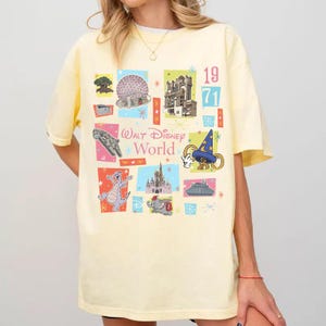 May include: A light yellow t-shirt featuring a colorful collage of Walt Disney World icons and the text "Walt Disney World" and "1971". The design includes images of the Spaceship Earth, Tower of Terror, and other park attractions.
