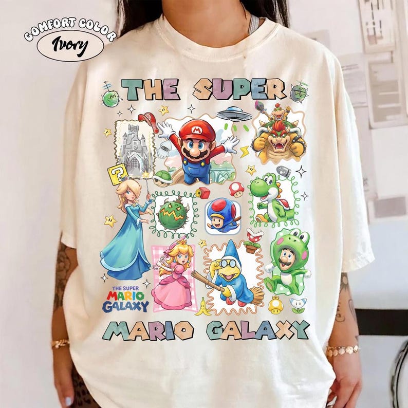 May include: Ivory-colored t-shirt featuring a colorful graphic design with characters and text from the Super Mario Galaxy video game. The text includes "THE SUPER MARIO GALAXY" in a playful font, along with various characters.