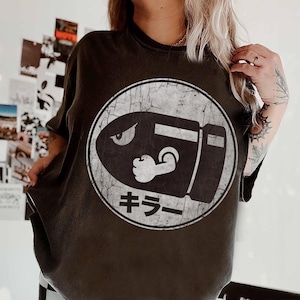 May include: Dark gray t-shirt featuring a distressed white graphic of a bullet with a muscular arm and the Japanese word "Killer" inside a circle. The shirt has a vintage, worn look.