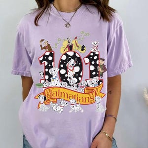 May include: A light purple t-shirt featuring a graphic of the Disney movie "101 Dalmatians." The design includes the number "101" with Dalmatian puppies and characters from the movie. The word "Dalmatians" is written on a banner.