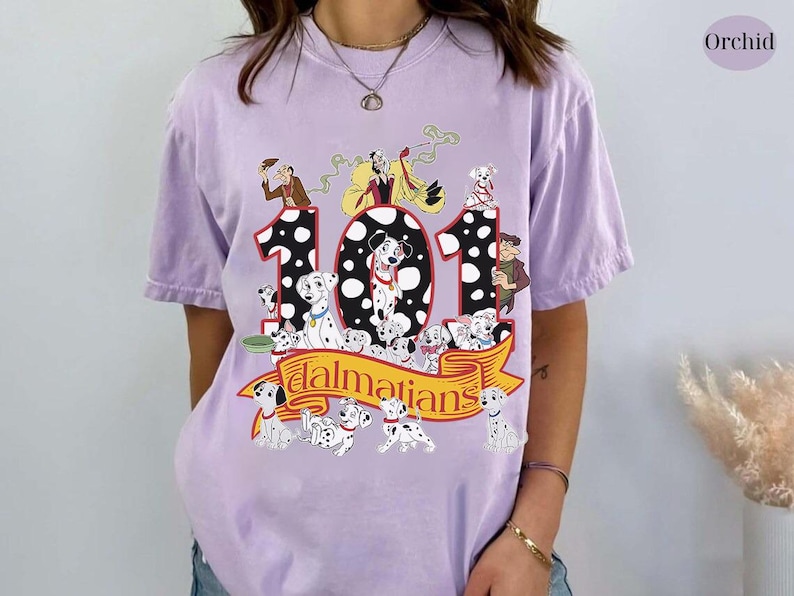 May include: A light purple t-shirt featuring a graphic of the Disney movie "101 Dalmatians." The design includes the number "101" with Dalmatian puppies and characters from the movie. The word "Dalmatians" is written on a banner.