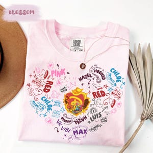 Puede incluir: Una camiseta de manga corta de color rosa claro con un gráfico central en forma de corazón compuesto por varios garabatos, textos como "JMJ", "CHLOE", "HAZEL", "LUIS", "MAX", "PINK", "RED", "VK VIBE", "RULE BREAKER" y un collar delicado.
