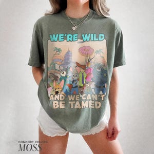 K&ouml;nnte beinhalten: Ein moosgr&uuml;nes Kurzarm-T-Shirt mit einem Grafikdruck verschiedener animierter Tierfiguren aus Zootopia. Der Text WE'RE WILD AND WE CAN'T BE TAMED ist in Hellblau und Beige &uuml;ber und unter der zentralen Illustration aufgedruckt.