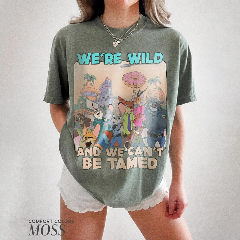 K&ouml;nnte beinhalten: Ein moosgr&uuml;nes Kurzarm-T-Shirt mit einem Grafikdruck verschiedener animierter Tierfiguren aus Zootopia. Der Text WE'RE WILD AND WE CAN'T BE TAMED ist in Hellblau und Beige &uuml;ber und unter der zentralen Illustration aufgedruckt.