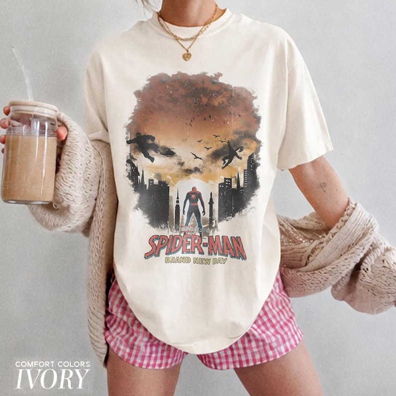 Pu&ograve; includere: Una t-shirt a maniche corte color avorio con una stampa grafica di Spider-Man in piedi in una silhouette cittadina contro un cielo al tramonto. Il design include il testo Spider-Man Brand New Day in lettere rosse e nere con uccelli.