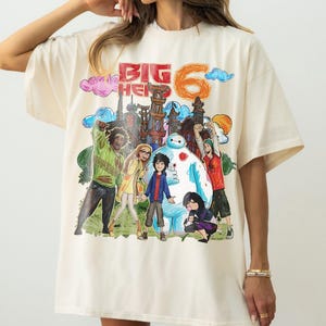 May include: Cream-colored t-shirt featuring a colorful illustration of characters and the text "BIG HERO 6". The graphic includes a cityscape, Baymax, and other figures in a cartoon style. The shirt has short sleeves.