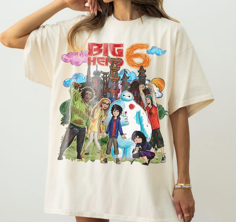 May include: Cream-colored t-shirt featuring a colorful illustration of characters and the text "BIG HERO 6". The graphic includes a cityscape, Baymax, and other figures in a cartoon style. The shirt has short sleeves.