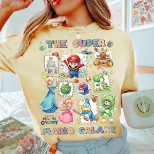 May include: Yellow t-shirt featuring a colorful graphic of characters from the Super Mario Galaxy video game. The design includes Mario, Princess Peach, Bowser, and other iconic figures, with the text "THE SUPER MARIO GALAXY".