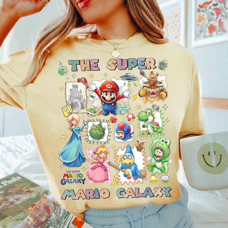 May include: Yellow t-shirt featuring a colorful graphic of characters from the Super Mario Galaxy video game. The design includes Mario, Princess Peach, Bowser, and other iconic figures, with the text "THE SUPER MARIO GALAXY".