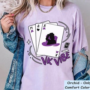 May include: A light purple t-shirt featuring a graphic of three playing cards with the letters M, A, and X, and a silhouette of a person wearing a top hat. The text VK VIBE is printed in purple below the cards, surrounded by small star and card icons.