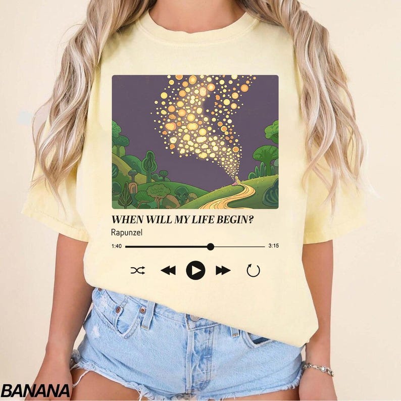 May include: A pale yellow t-shirt featuring a graphic print of a music player interface. The design displays a scene of floating lanterns over a landscape, with the text "WHEN WILL MY LIFE BEGIN?" and "Rapunzel" above a playback progress bar.