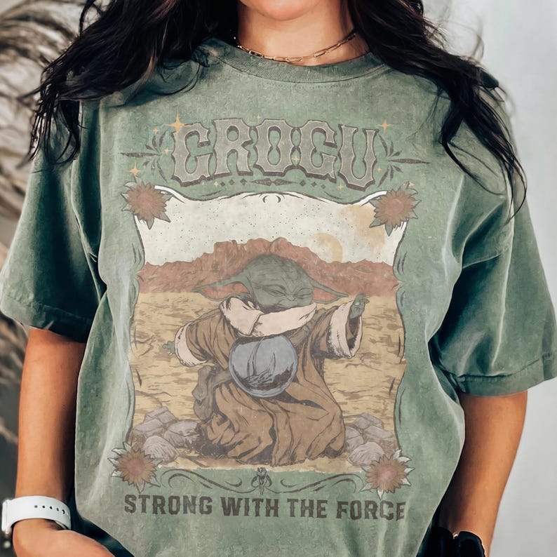May include: Green t-shirt featuring a graphic of Grogu from The Mandalorian. The design includes the text "GROGU" above the character and "STRONG WITH THE FORCE" below. The image has a vintage, distressed look.