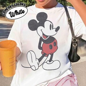Peut inclure: T-shirt blanc avec un dessin de Mickey Mouse en noir, blanc et rouge. Le t-shirt porte les mots "COMFORT COLORS" et "White". Le t-shirt est un v&ecirc;tement d&eacute;contract&eacute;.