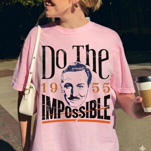 Pu&ograve; includere: Una maglietta rosa chiaro con la scritta "Do The Impossible" in nero, con l'anno 1955 in marrone. La maglietta presenta un ritratto di un uomo con i baffi. Un capo casual.