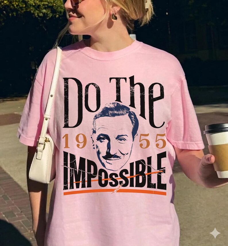 Pu&ograve; includere: Una maglietta rosa chiaro con la scritta "Do The Impossible" in nero, con l'anno 1955 in marrone. La maglietta presenta un ritratto di un uomo con i baffi. Un capo casual.