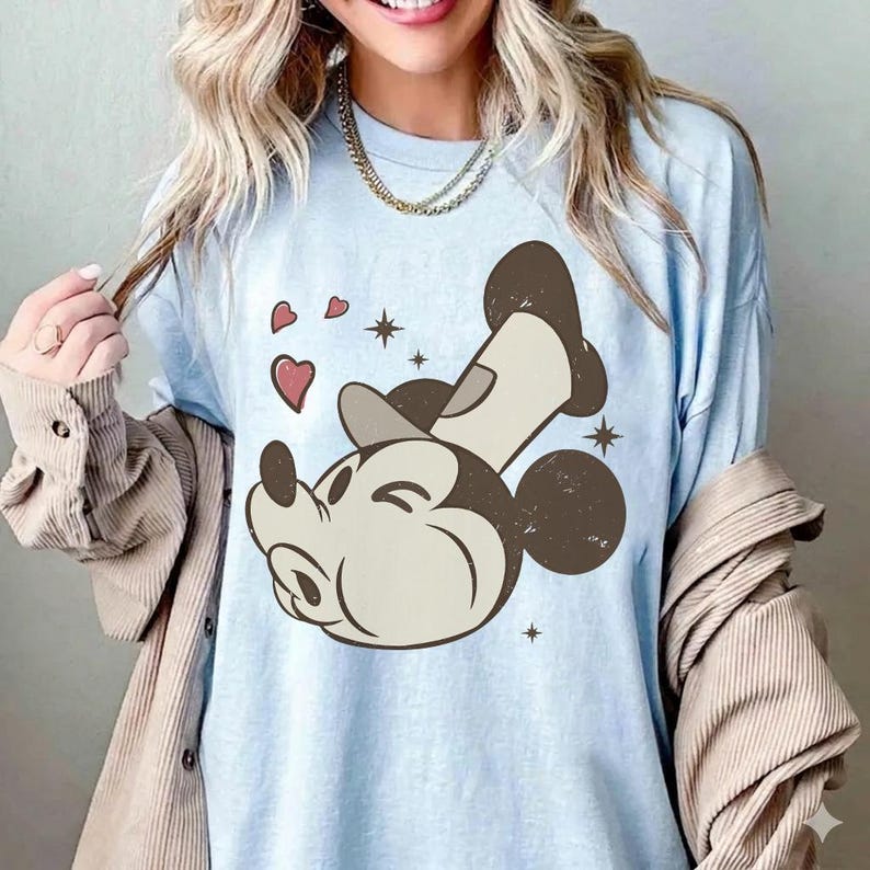 May include: Light blue t-shirt featuring a cartoon Mickey Mouse design. The graphic shows Mickey with a wink, surrounded by hearts and stars. The shirt is a relaxed fit, with a crew neck and short sleeves.