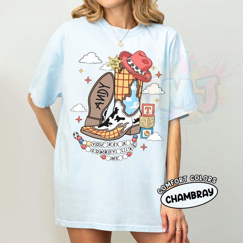 May include: A light blue t-shirt featuring a graphic of a cowboy boot with a cow print pattern, a red cowboy hat, a sheriff badge, alphabet blocks, and a string of beads. The text reads ANDY, INFINITY AND BEYOND, and YOU'RE A COWBOY LIKE ME.