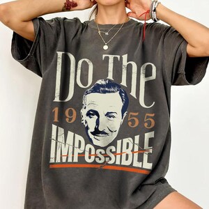 Pu&ograve; includere: T-shirt grigio scuro con la scritta "Do The Impossible" e un ritratto di un uomo con i baffi. Anche i numeri "1955" sono stampati sulla maglietta. La maglietta ha un aspetto vintage e usurato.