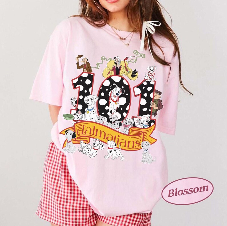 May include: A light pink t-shirt featuring a graphic of the number "101" with Dalmatian characters and the word "Dalmatians" on a banner. The shirt has short sleeves and a round neckline. The shirt is paired with red and white checkered shorts.