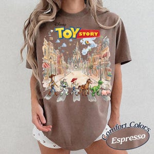 May include: Espresso-colored t-shirt featuring a Disney Toy Story graphic. The graphic depicts the Toy Story characters crossing a street with a castle in the background. The shirt is labeled "Comfort Colors".