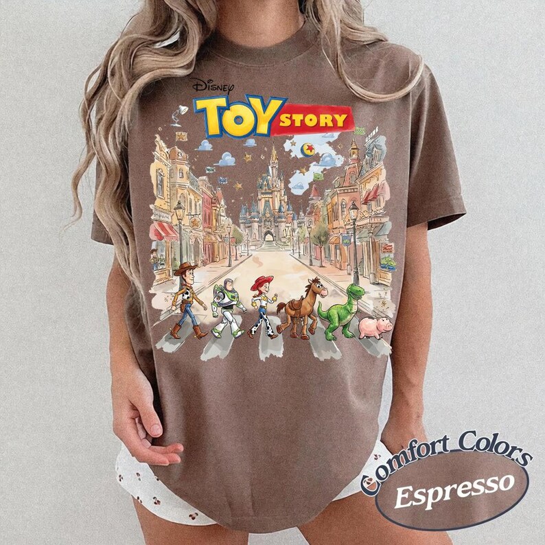 May include: Espresso-colored t-shirt featuring a Disney Toy Story graphic. The graphic depicts the Toy Story characters crossing a street with a castle in the background. The shirt is labeled "Comfort Colors".