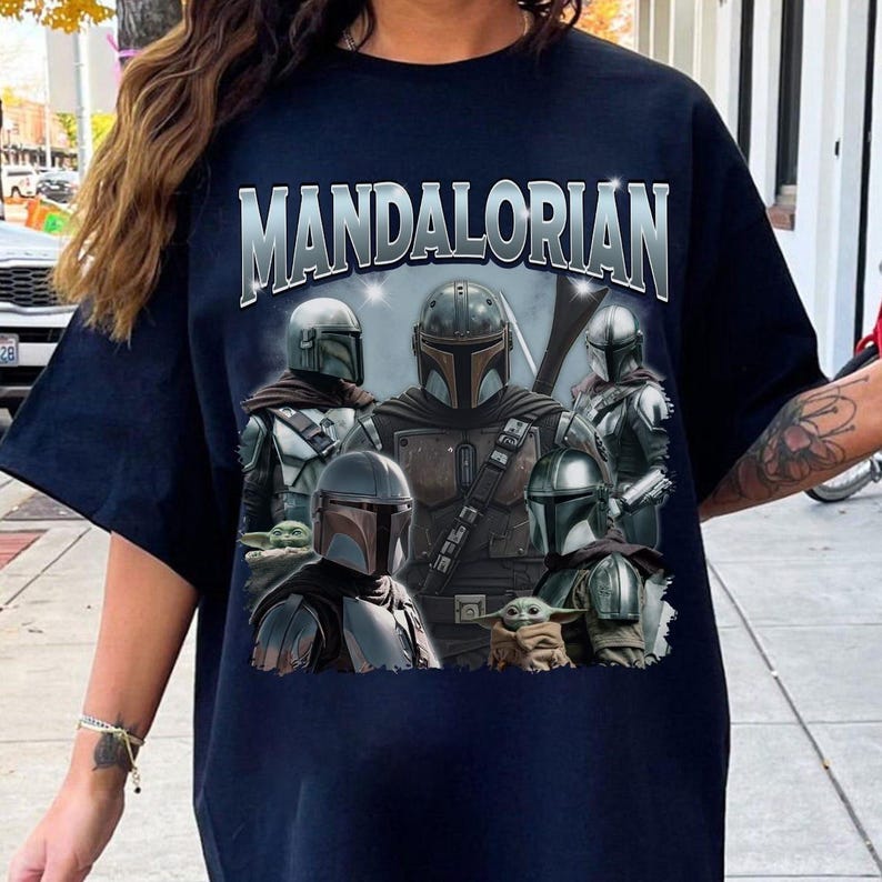 May include: Navy blue t-shirt featuring the word "MANDALORIAN" in silver lettering above a graphic of several Mandalorian characters and Grogu. The design includes various helmets and armor, with a light blue and gray background.