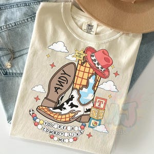 May include: A cream-colored t-shirt featuring a graphic of a cowboy boot with a cow-print pattern, a red hat, a sheriff star labeled "Infinity and Beyond," alphabet blocks, and a beaded necklace with the text "You're a cowboy like me."