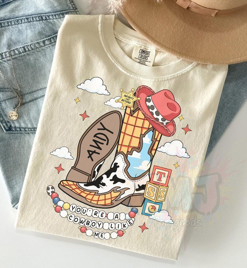 May include: A cream-colored t-shirt featuring a graphic of a cowboy boot with a cow-print pattern, a red hat, a sheriff star labeled "Infinity and Beyond," alphabet blocks, and a beaded necklace with the text "You're a cowboy like me."