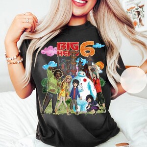 May include: Black t-shirt featuring a colorful graphic of characters from the animated movie "Big Hero 6." The design includes the text "BIG HERO 6" in red and orange, with illustrations of the main characters and a cityscape background.