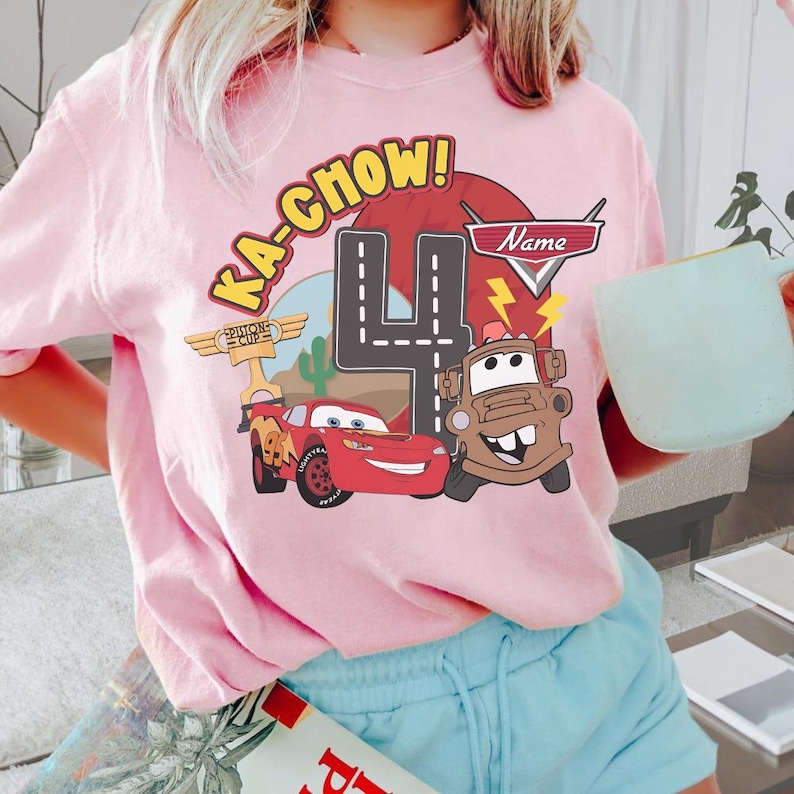 May include: Pink t-shirt featuring a graphic of Lightning McQueen and Mater from the movie Cars, with the text "KA-CHOW!" and the number "4". The design includes a road, cactus, and a "Name" banner.