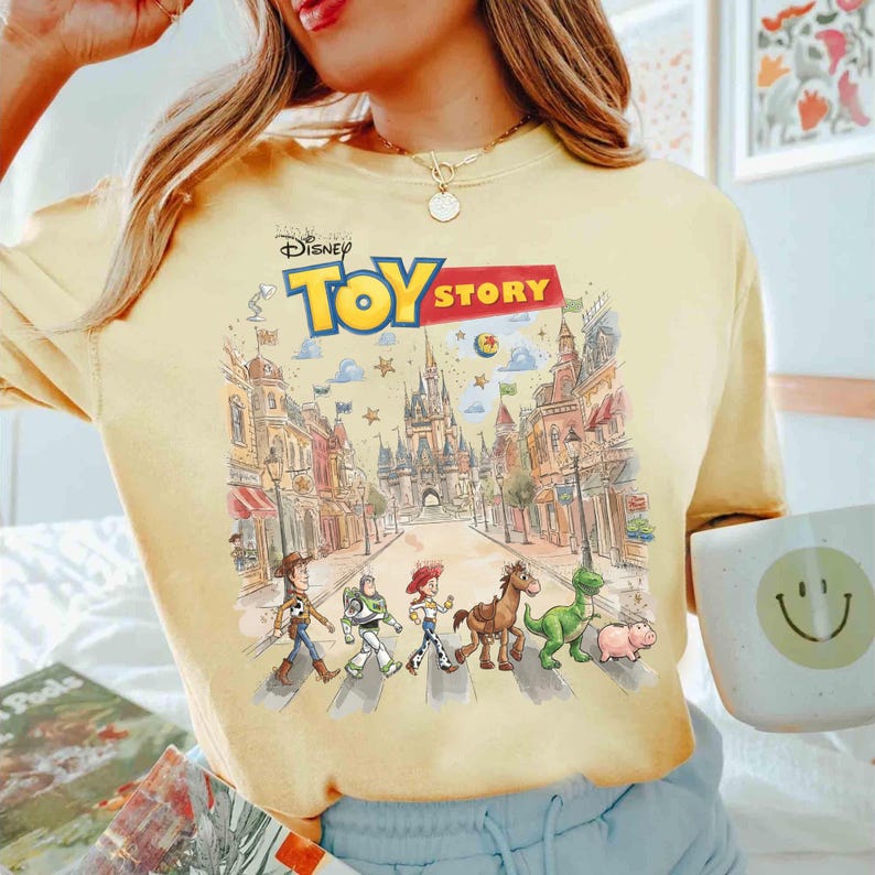 May include: Yellow t-shirt featuring a colorful graphic of Toy Story characters crossing a street in front of a castle. The Disney logo and "TOY STORY" text are at the top. The shirt is a light yellow color.