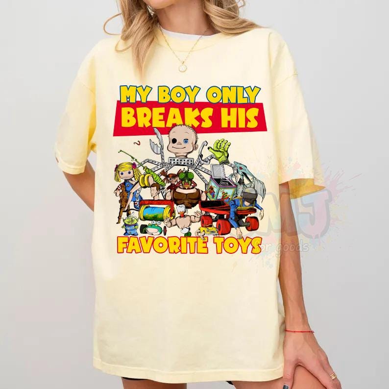 May include: A pale yellow short-sleeve t-shirt featuring a graphic print of various toys and a doll head. The text reads MY BOY ONLY BREAKS HIS FAVORITE TOYS in bold yellow and red lettering. The design includes a jack-in-the-box and other figures.