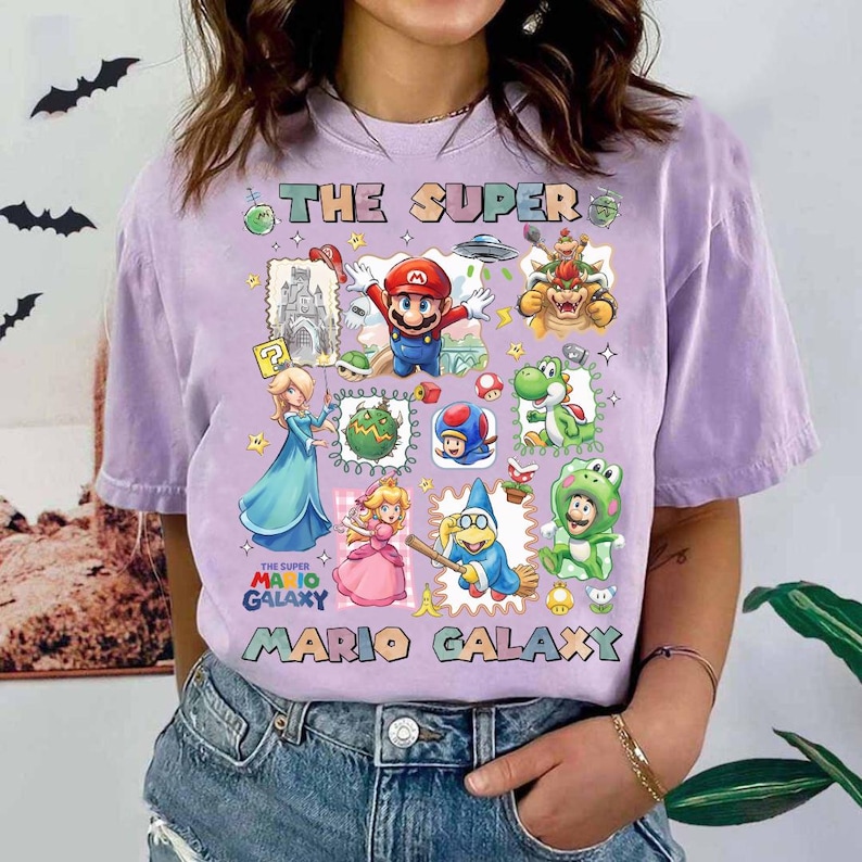 May include: A lilac-colored t-shirt featuring a colorful graphic of characters from the Super Mario Galaxy video game. The design includes Mario, Princess Peach, Yoshi, and Bowser, with the text "THE SUPER MARIO GALAXY".