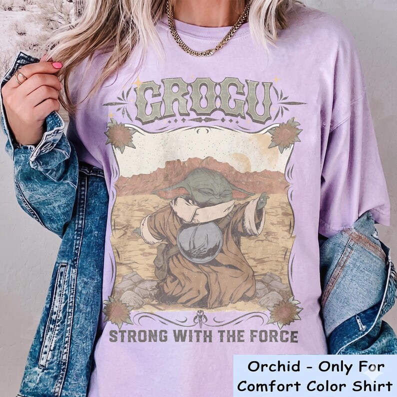 May include: Orchid-colored t-shirt featuring a graphic of Grogu, with the text "GROGU" above and "STRONG WITH THE FORCE" below. The design includes a desert landscape and floral accents. The shirt is a Comfort Color shirt.