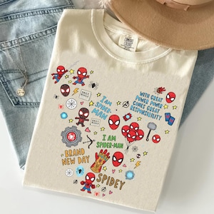 Puede incluir: Camiseta color crema con un colorido gr&aacute;fico de Spider-Man. El dise&ntilde;o incluye varias ilustraciones de Spider-Man, el texto "I am Spider-Man" y otros s&iacute;mbolos relacionados. La camiseta est&aacute; doblada sobre un par de jeans.